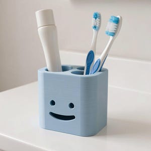 Puede incluir: Un porta cepillos de dientes azul claro con un dise&ntilde;o de cara sonriente. El soporte contiene dos cepillos de dientes con detalles azules y un tubo de pasta de dientes. El soporte est&aacute; hecho de un material texturizado y se encuentra sobre una superficie blanca.