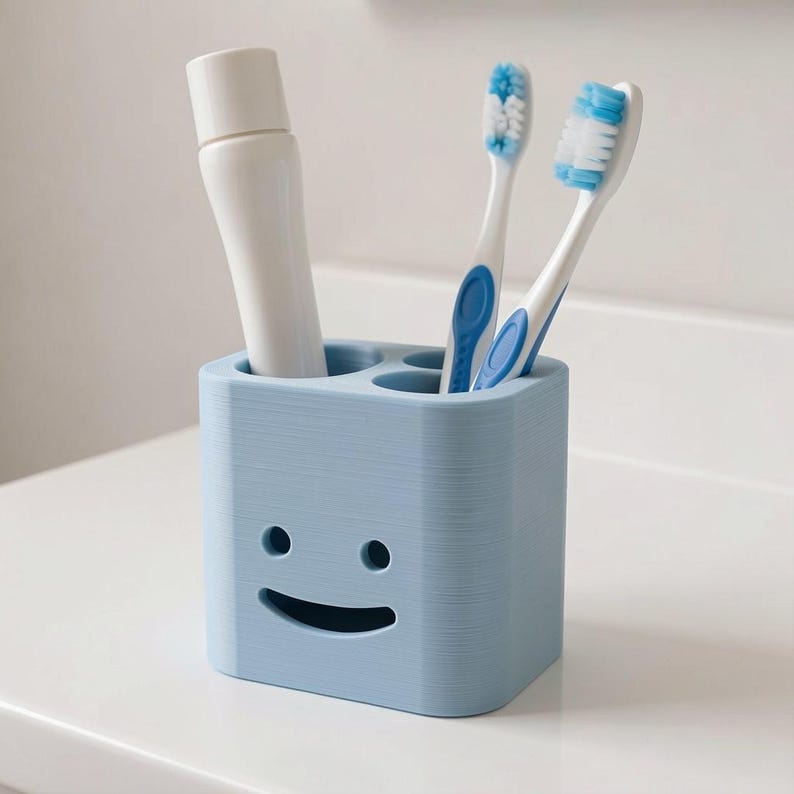 Puede incluir: Un porta cepillos de dientes azul claro con un dise&ntilde;o de cara sonriente. El soporte contiene dos cepillos de dientes con detalles azules y un tubo de pasta de dientes. El soporte est&aacute; hecho de un material texturizado y se encuentra sobre una superficie blanca.