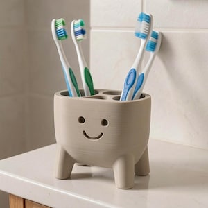 Pu&ograve; includere: Un portaspazzolini beige con un design a faccina sorridente, che contiene diversi spazzolini con setole blu e verdi. Il supporto ha una forma rettangolare arrotondata ed &egrave; sostenuto da tre piccole gambe.