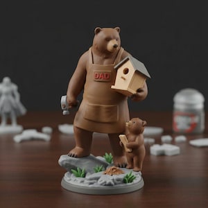 K&ouml;nnte beinhalten: Eine braune B&auml;renfigur mit einer braunen Sch&uuml;rze mit dem Wort "DAD" und einem Hammer und einem Vogelhaus. Ein kleineres B&auml;renjunges h&auml;lt ein St&uuml;ck Holz. Die Figur steht auf einem grauen Sockel mit gr&uuml;nem Gras und Felsen.