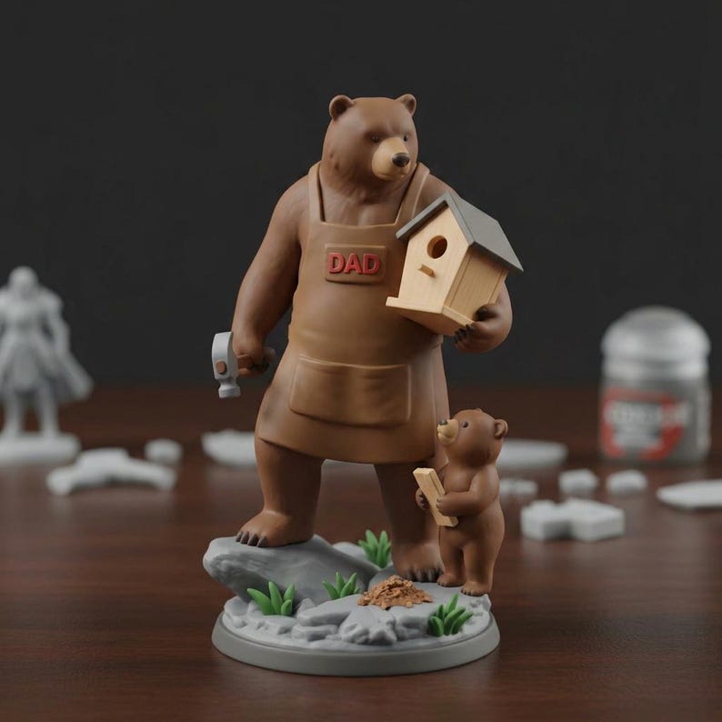 K&ouml;nnte beinhalten: Eine braune B&auml;renfigur mit einer braunen Sch&uuml;rze mit dem Wort "DAD" und einem Hammer und einem Vogelhaus. Ein kleineres B&auml;renjunges h&auml;lt ein St&uuml;ck Holz. Die Figur steht auf einem grauen Sockel mit gr&uuml;nem Gras und Felsen.