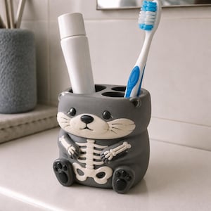 May include: A gray ceramic toothbrush holder shaped like an otter with a white skeleton graphic on its chest. The holder contains a white tube of toothpaste and a blue and white toothbrush. The otter has black eyes, a black nose, and paw details.