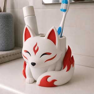May include: A white and red fox-shaped toothbrush holder with a matte finish. The fox has closed eyes, a black nose, and red flame-like markings on its forehead, ears, and large curled tail. It contains a white tube and a blue and white toothbrush.