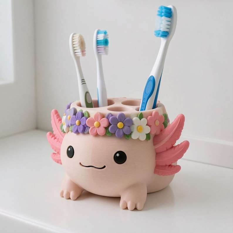May include: A light pink axolotl-shaped toothbrush holder featuring a crown of colorful purple, pink, and white flowers. The holder has three circular openings containing three toothbrushes with white, blue, and green handles.