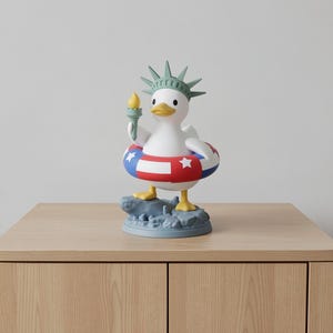 May include: A whimsical figurine of a white duck, adorned with a Statue of Liberty crown and holding a torch. It wears a red, white, and blue inflatable ring. The duck stands on a blue base, evoking a patriotic theme.