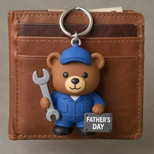 May include: A brown bear keychain wearing a blue cap and blue work uniform, holding a silver wrench in one hand and a gray sign that reads FATHER'S DAY in the other, attached to a silver metal ring resting on a brown leather bifold wallet.