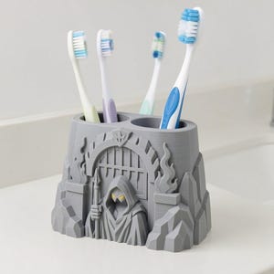 Puede incluir: Un soporte para cepillos de dientes gris con una figura esculpida con capucha que sostiene una lanza frente a un arco con rejas. El soporte tiene dos aberturas circulares y contiene cuatro cepillos de dientes con cerdas y mangos blancos, azules y verdes.