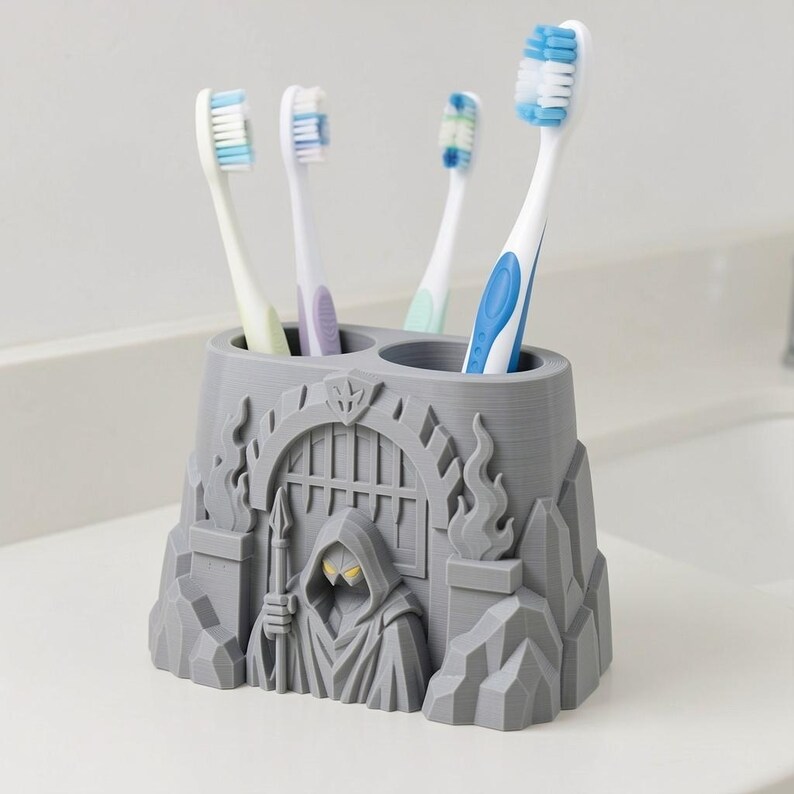 Puede incluir: Un soporte para cepillos de dientes gris con una figura esculpida con capucha que sostiene una lanza frente a un arco con rejas. El soporte tiene dos aberturas circulares y contiene cuatro cepillos de dientes con cerdas y mangos blancos, azules y verdes.