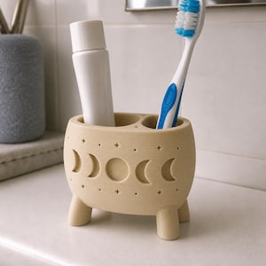 May include: A beige toothbrush holder with three short legs, featuring a debossed moon phase cycle and small star patterns. The holder contains a white tube of toothpaste and a blue and white toothbrush with blue bristles standing upright.