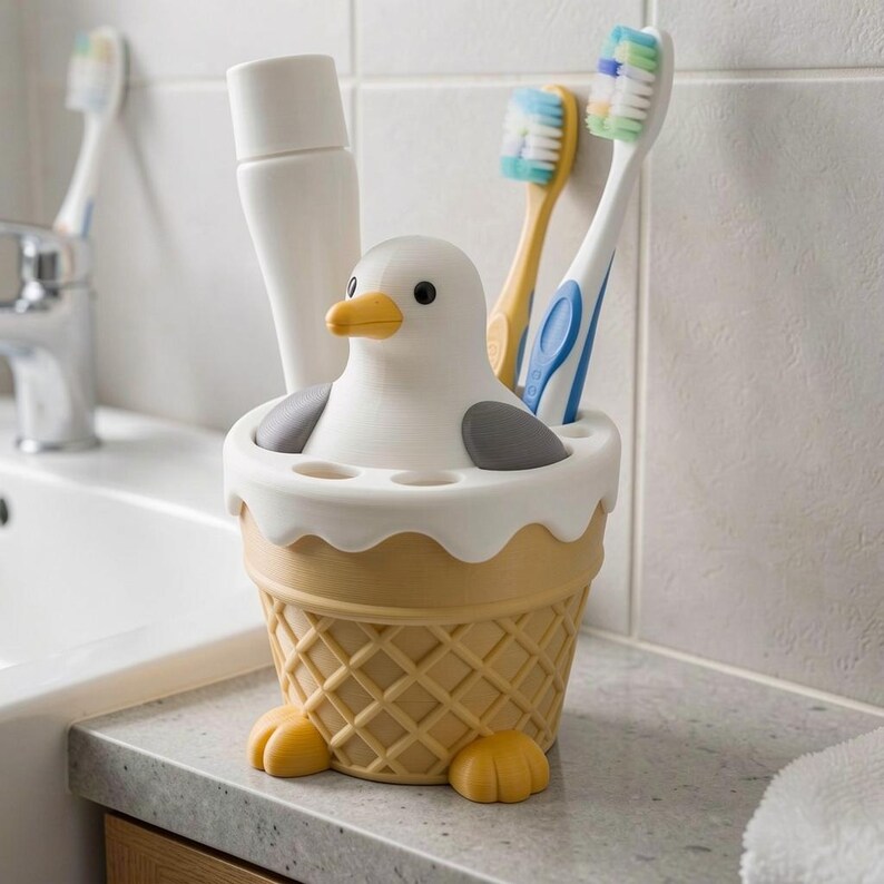 May include: A white and yellow seagull-shaped toothbrush holder designed like an ice cream cone with a waffle-textured base. The holder features a white bird with grey wings and a yellow beak, containing a white tube and two toothbrushes.