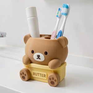 Puede incluir: Un soporte para cepillos de dientes con forma de oso marr&oacute;n, cabeza de color marr&oacute;n claro y base rectangular amarilla con la palabra BUTTER. El soporte tiene dos peque&ntilde;as patas en la parte delantera, huellas en los pies y dos cepillos.