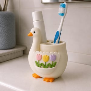 Pu&ograve; includere: Un portaspazzolino a forma di anatra color crema con piedi e becco arancioni. L'anatra presenta una tasca decorativa sulla parte anteriore che mostra tre tulipani color pastello rosa, gialli e viola con steli e foglie di colore verde.