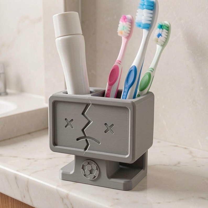 Puede incluir: Un soporte rectangular gris para cepillos de dientes con un rostro estilizado de ojos en forma de X y boca agrietada. El soporte contiene un tubo blanco y tres cepillos de dientes con cerdas rosas, azules y verdes. Un icono de bal&oacute;n de f&uacute;tbol en la base.