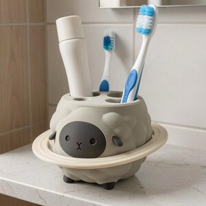 May include: A light gray sheep-shaped toothbrush holder with a dark gray face and a cream-colored ring around its middle. The holder features multiple circular openings on top, currently containing a white tube and two toothbrushes with blue bristles.