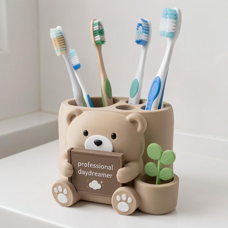 May include: A beige bear-shaped toothbrush holder with four circular slots holding four toothbrushes. The bear holds a brown rectangular sign that reads professional daydreamer with a small cloud icon. A small green plant sits to the right.