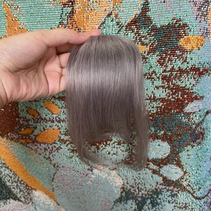 May include: A clip-in hair extension piece featuring straight, ash-toned light brown hair with a soft, cool-toned finish. The hair is styled with a blunt, straight-cut fringe that frames the face, showcasing a smooth texture and natural-looking sheen.