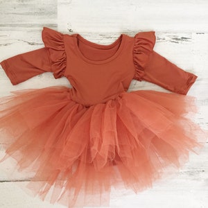 May include: A terracotta-colored long-sleeved dress with a tulle skirt. The bodice has ruffled sleeves and a round neckline. The skirt is made of layers of soft, peach-colored tulle, creating a full, flowing effect. The dress is designed for a child.