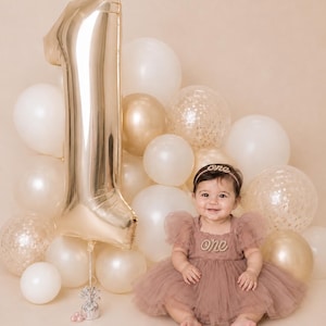 Peut inclure: Une robe en tulle rose poudr&eacute; avec une application perl&eacute;e 'one', un bandeau assorti 'one' et un grand ballon en aluminium dor&eacute; en forme de chiffre un, entour&eacute; d'un groupe de ballons blancs, dor&eacute;s et transparents remplis de confettis.