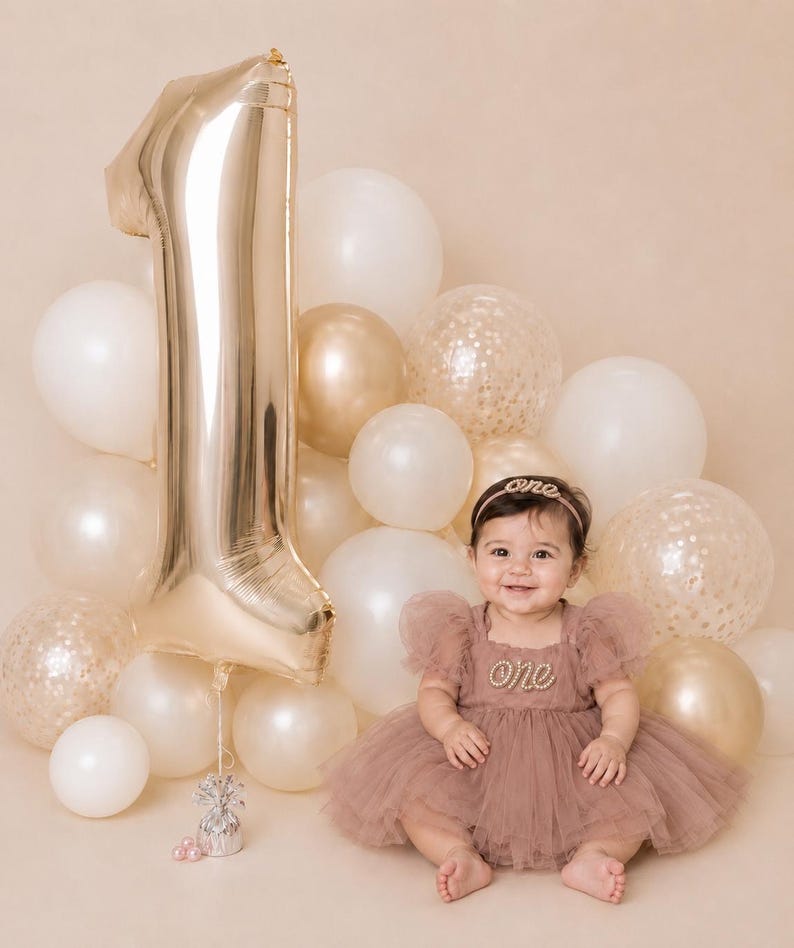 Peut inclure: Une robe en tulle rose poudr&eacute; avec une application perl&eacute;e 'one', un bandeau assorti 'one' et un grand ballon en aluminium dor&eacute; en forme de chiffre un, entour&eacute; d'un groupe de ballons blancs, dor&eacute;s et transparents remplis de confettis.