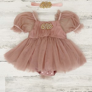 Peut inclure: Une barboteuse en tulle rose poudr&eacute; avec des manches bouffantes, une encolure carr&eacute;e et un &eacute;cusson perl&eacute; sur la poitrine portant l'inscription "one". Un bandeau assorti avec une d&eacute;coration perl&eacute;e "one" accompagne la tenue.