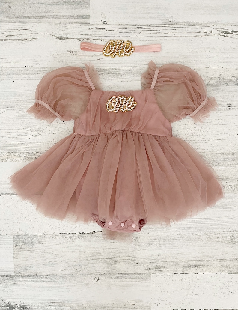 Peut inclure: Une barboteuse en tulle rose poudr&eacute; avec des manches bouffantes, une encolure carr&eacute;e et un &eacute;cusson perl&eacute; sur la poitrine portant l'inscription "one". Un bandeau assorti avec une d&eacute;coration perl&eacute;e "one" accompagne la tenue.