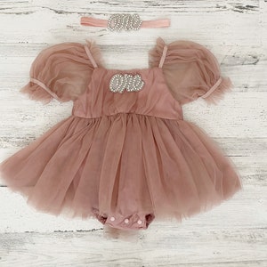 Peut inclure: Une barboteuse en tulle rose poudr&eacute; avec des manches bouffantes courtes, une encolure carr&eacute;e et une fermeture &agrave; boutons-pression en bas. La poitrine affiche le mot "one" en strass argent&eacute;s. Un bandeau assorti avec le mot "one" est inclus.