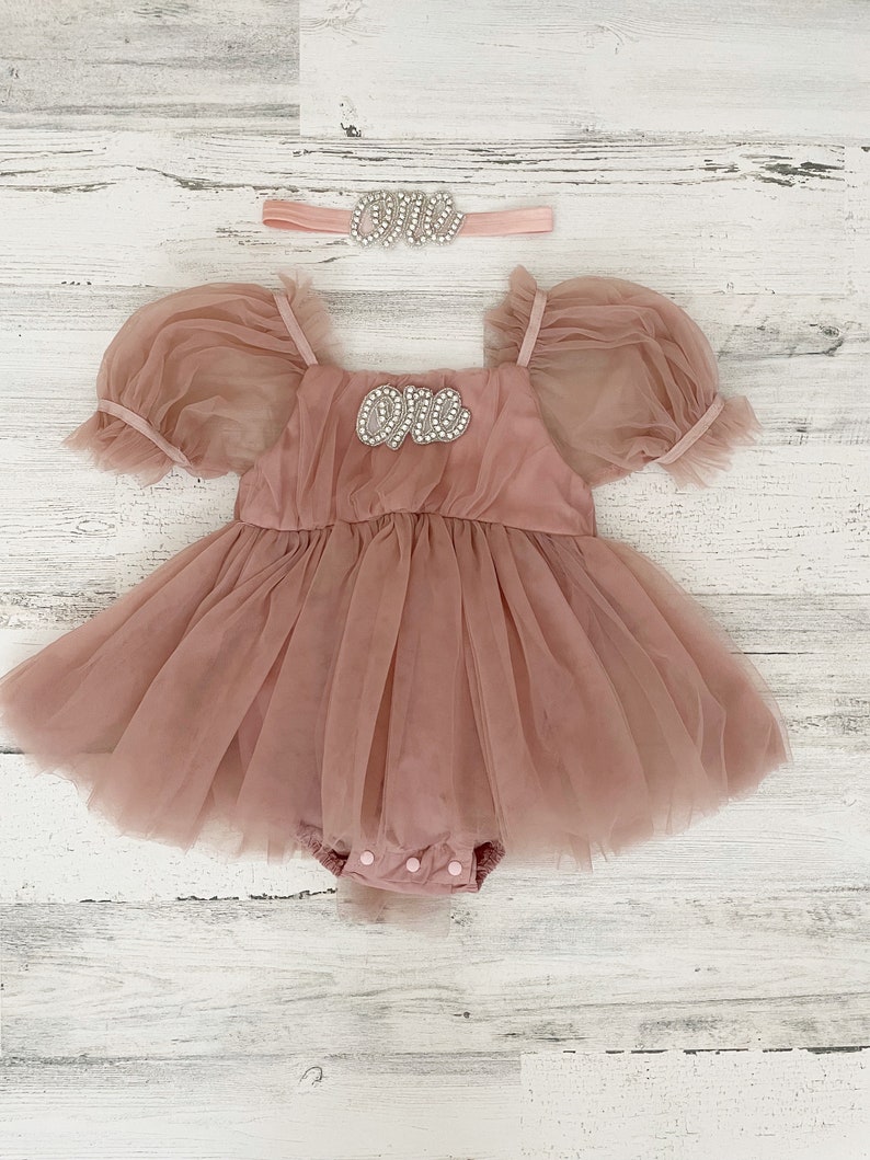 Peut inclure: Une barboteuse en tulle rose poudr&eacute; avec des manches bouffantes courtes, une encolure carr&eacute;e et une fermeture &agrave; boutons-pression en bas. La poitrine affiche le mot "one" en strass argent&eacute;s. Un bandeau assorti avec le mot "one" est inclus.