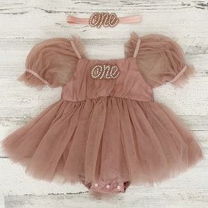Peut inclure: Une barboteuse en tulle rose poudr&eacute; avec des manches bouffantes, une encolure carr&eacute;e et un &eacute;cusson perl&eacute; sur la poitrine portant l'inscription "one" en &eacute;criture cursive. Un bandeau assorti avec un &eacute;cusson "one" perl&eacute; est inclus.