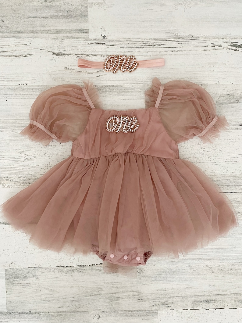 Peut inclure: Une barboteuse en tulle rose poudr&eacute; avec des manches bouffantes, une encolure carr&eacute;e et un &eacute;cusson perl&eacute; sur la poitrine portant l'inscription "one" en &eacute;criture cursive. Un bandeau assorti avec un &eacute;cusson "one" perl&eacute; est inclus.