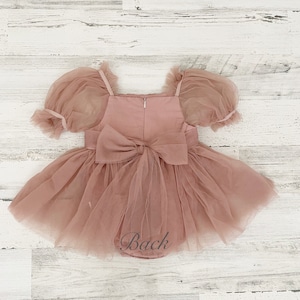 Peut inclure: Une barboteuse rose poudr&eacute; avec des manches bouffantes transparentes, une encolure carr&eacute;e et une jupe en tulle superpos&eacute;e. Le v&ecirc;tement pr&eacute;sente un grand n&oelig;ud en tissu au centre du dos, une fermeture &eacute;clair verticale et le mot Back en gris.