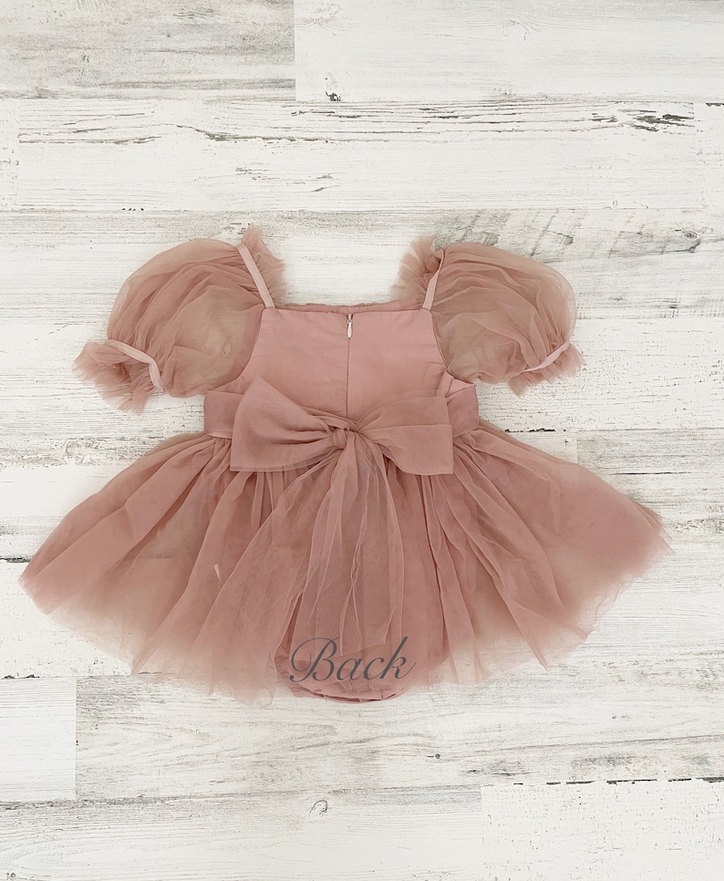 Peut inclure: Une barboteuse rose poudr&eacute; avec des manches bouffantes transparentes, une encolure carr&eacute;e et une jupe en tulle superpos&eacute;e. Le v&ecirc;tement pr&eacute;sente un grand n&oelig;ud en tissu au centre du dos, une fermeture &eacute;clair verticale et le mot Back en gris.