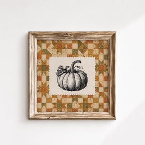 May include: A square wall art piece in a weathered brown wooden frame. The centre features a cream-coloured fabric patch with a black ink illustration of a pumpkin. The background is a patchwork quilt pattern in shades of orange, green, and tan.