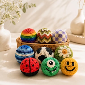 May include: A collection of nine round, crocheted yarn balls in a woven basket. Designs include a rainbow, chevron pattern, purple checkerboard, blue stripes, cow print, white daisy on green, red ladybug, green one-eyed monster, and yellow smiley face.