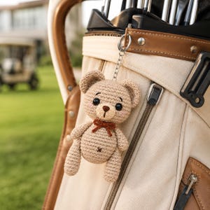 Puede incluir: Un llavero de oso de peluche tejido a ganchillo en color beige con una pequeña pajarita marrón, ojos de plástico negro y nariz bordada. El oso está sujeto a una cadena y anilla de metal plateado, colgado de una bolsa de golf clara con detalles en cuero marrón.