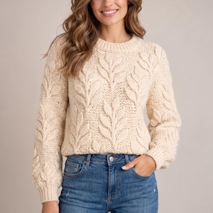 May include: A cream-colored long-sleeve sweater featuring a raised, textured leaf pattern across the front and sleeves. The garment has a ribbed crew neckline and ribbed cuffs, paired with blue denim jeans with a visible metal button.