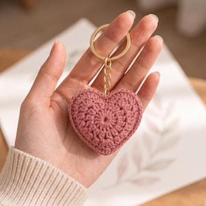 Puede incluir: Un llavero en forma de corazón de color rosa empolvado hecho de hilo de ganchillo. El corazón presenta un patrón de puntos radiales texturizados y está unido a una anilla de metal dorado mediante una cadena corta de eslabones dorados.
