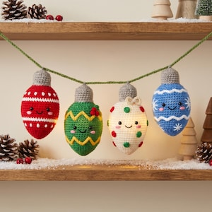 May include: A garland featuring four crocheted Christmas light bulb ornaments in red, green, white, and blue, each with a grey cap, embroidered smiling faces, and pink cheeks, suspended from a green yarn string against a neutral background.
