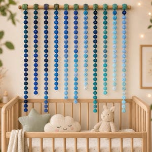 May include: A wooden crib with a decorative mobile of blue crochet balls hanging above. The mobile features a gradient of blue hues, from deep indigo to light turquoise. A star and cloud pillow and a stuffed bunny are in the crib.