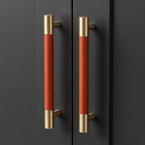 May include: Two vertical cabinet pulls featuring a central cylindrical grip wrapped in textured burnt orange material, accented by brushed gold metal end caps and mounting posts, attached to a dark charcoal gray cabinet door surface.