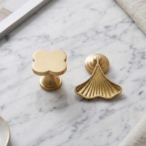 May include: Two gold-toned metal cabinet knobs. One knob features a four-lobed clover shape with a smooth, matte finish. The second knob is shaped like a fan or ginkgo leaf with vertical ridges and a circular base, both in a brushed gold finish.