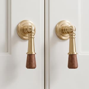 May include: Two cabinet pulls featuring a circular brushed brass base and a vertical, tapered metal arm. The lower section of each handle consists of a cylindrical, dark brown wood grip with a visible grain texture, mounted on white doors.