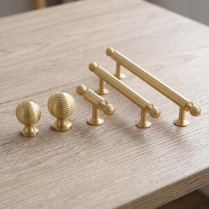 May include: A collection of five gold-toned metal cabinet hardware pieces, including two spherical knobs with horizontal ribbed detailing and three bar-style pulls of varying lengths, each featuring matching ribbed end caps.