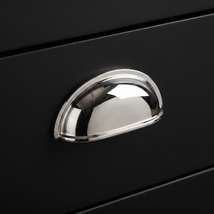 Peut inclure: Une poignée de tirage en forme de coquille en chrome poli avec un design semi-circulaire lisse, fixée sur une façade de tiroir noir mat. La finition métallique reflète la lumière, soulignant la forme incurvée et la fixation horizontale.