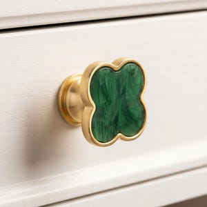 May include: A clover-shaped drawer knob featuring a brushed gold metal frame and a marbled dark green resin inlay. The hardware has a rounded base and a four-lobed design, providing a decorative accent for furniture cabinetry and drawers.