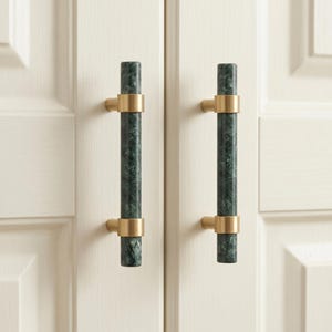 May include: Two vertical cabinet pulls featuring cylindrical dark green marble shafts with visible white veining. Each handle is secured by two brushed gold metal mounts at the top and bottom, attached to a light cream-colored paneled cabinet door.