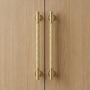 Puede incluir: Dos tiradores de armario verticales de latón con textura acanalada y extremos cilíndricos lisos. Los tiradores están montados en puertas de armario de madera de roble claro, con un patrón de veta vertical y una unión recta.
