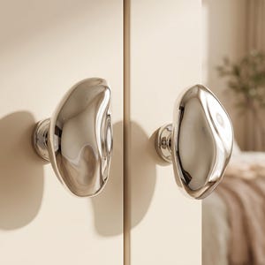 May include: Two polished silver-toned cabinet knobs with an irregular, organic, pebble-like shape. Each knob features a smooth, reflective metallic surface and is mounted on a circular base attached to a light cream-coloured cabinet door panel.