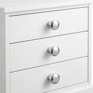 May include: A white wooden nightstand featuring three horizontal drawers, each equipped with a single polished chrome knob. The knobs have a scalloped, flower-like edge design. The furniture piece has a clean, smooth finish and a rectangular frame.