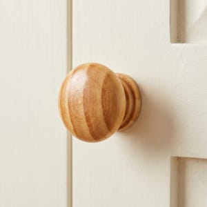 May include: A round, light brown wooden cabinet knob with a smooth, polished finish and visible natural wood grain patterns. The knob features a spherical head attached to a smaller, cylindrical base, mounted onto a flat, off-white surface.