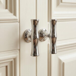 May include: A pair of polished chrome cabinet handles with a vertical, cylindrical, and slightly contoured design. Each handle is mounted on a circular base plate, attached to a cream-colored cabinet door with a recessed panel frame detail.